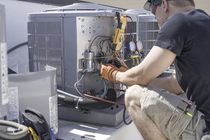 Air conditioner repair in Manasquan being done on condenser by man in black shirt and tan shorts wearing work gloves and safety goggles.