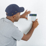person in blue baseball cap with both hands on a thermostat setting it to proper settings to avoid air conditioner repair in Manasquan